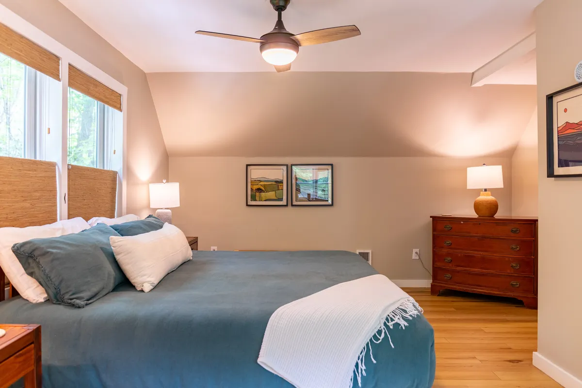 A cozy bedroom with a bed featuring teal bedding, a wooden dresser, decorative artwork, and soft lamps on nightstands, all bathed in natural light. The serene mood is perfect for relaxing or quiet evenings.