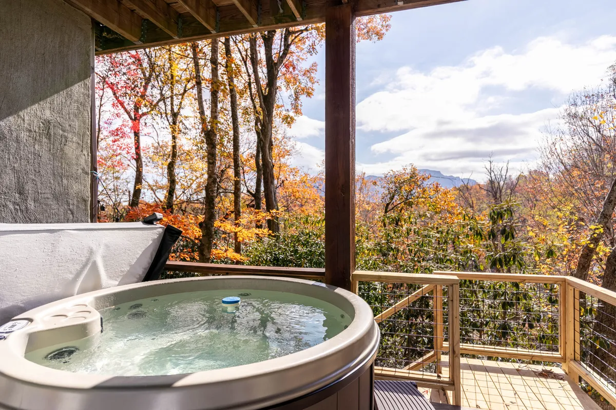 A round hot tub sits on a covered deck, surrounded by beautiful autumn trees and a mountain backdrop. The scene exudes relaxation, with the tub inviting guests to unwind while soaking in scenic views of nature.