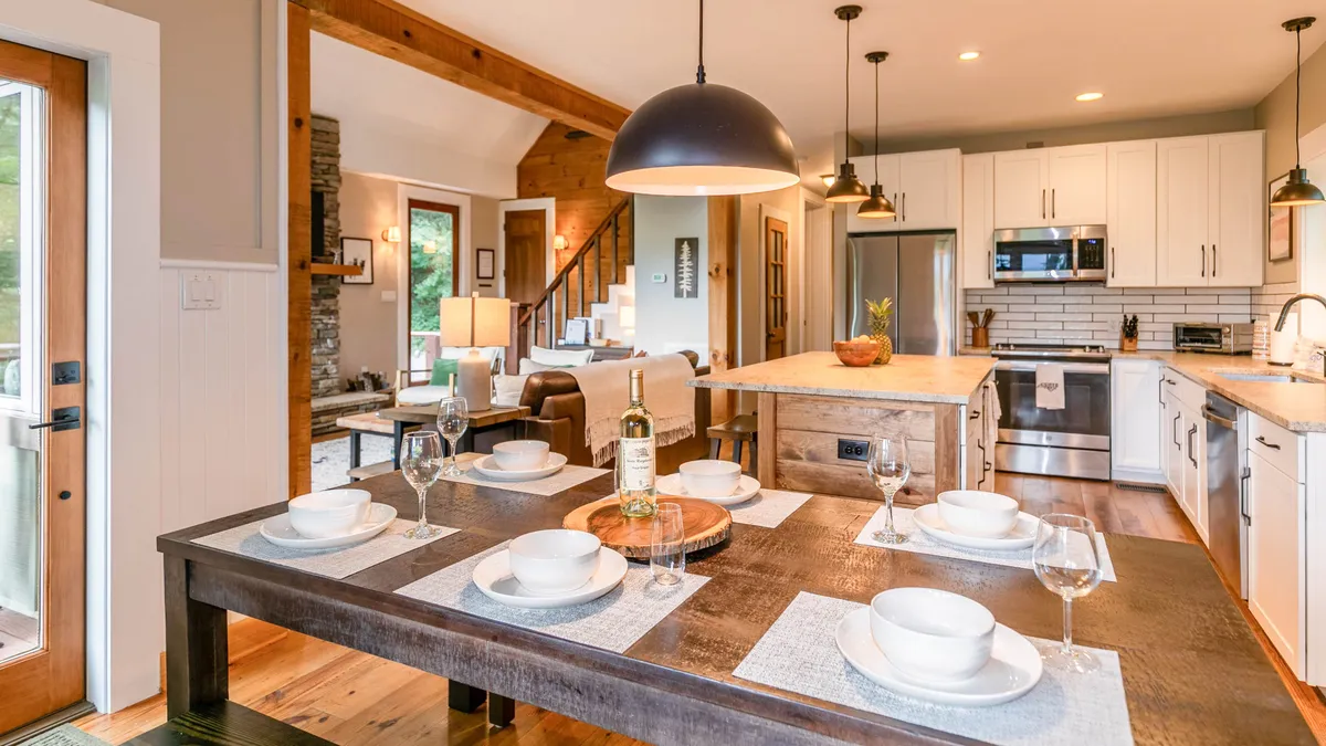 An open-concept interior with a wooden-beamed ceiling, a dining table set for a meal, and a kitchen island in the background. The layout allows seamless flow between living, dining, and kitchen areas, reinforcing the sense of togetherness.