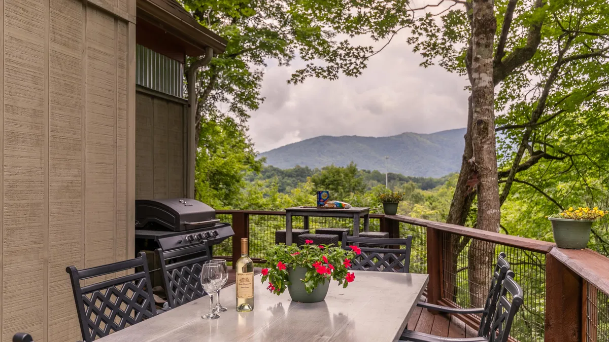 A tranquil mountain deck featuring a dining table set with wine and glasses, a grill, and scenic views of the surrounding peaks and flower pots. The space invites outdoor dining and relaxation in nature.