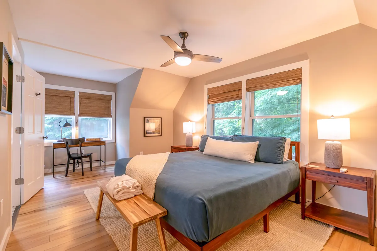 A tranquil bedroom with a plush bed, wood nightstand, and a window letting in natural light. A small desk and lamp provide a serene corner for reading or morning routines, emphasizing comfort and relaxation.