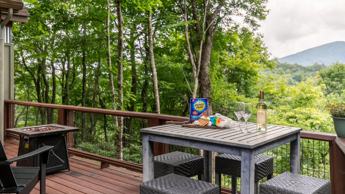 A welcoming outdoor dining area with snacks and wine set on a table, a fire pit nearby, and panoramic mountain views. The surrounding greenery and mountain vista create a tranquil, inviting space.