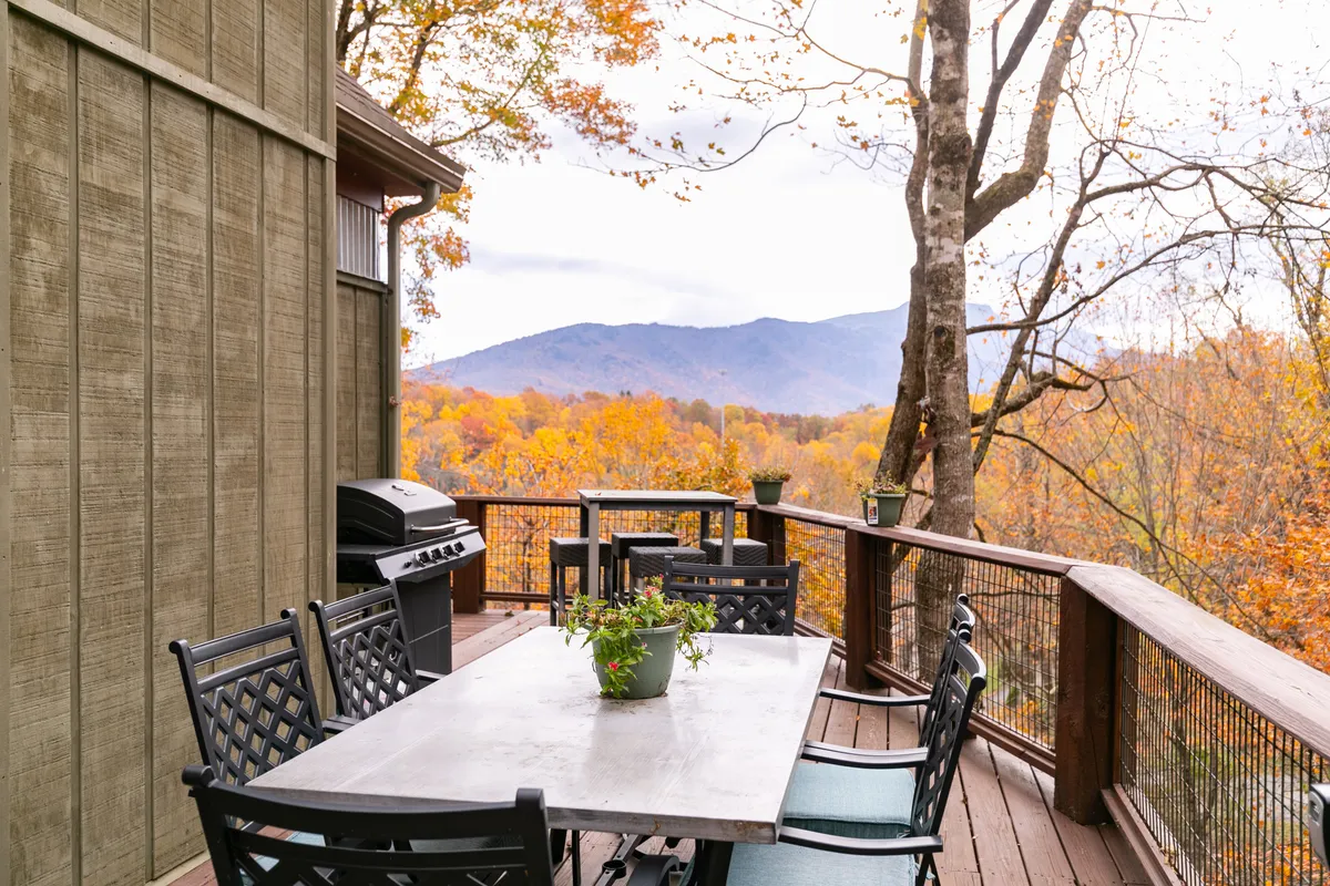 An inviting outdoor dining area with a table set for a meal, a grill, and a panoramic view of rolling, tree-covered mountains. The fall foliage and golden light set a warm, communal mood for al fresco dining or cookouts.
