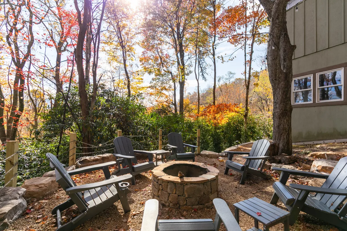 The photo showcases a fire pit encircled by Adirondack chairs in a clearing amidst colorful fall foliage and tall trees. The arrangement encourages communal gatherings, ideal for storytelling, making s'mores, or simply enjoying nature.