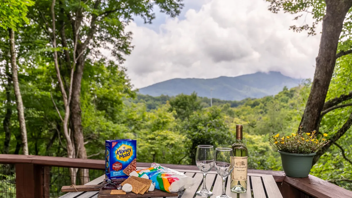 The image showcases an inviting wooden outdoor table set with wine glasses and snacks, overlooking a breathtaking mountain view at sunset. The scene captures a sense of relaxation and camaraderie, ideal for sharing meals and laughter with friends or family while soaking in the stunning scenery of Seven Devils, North Carolina.