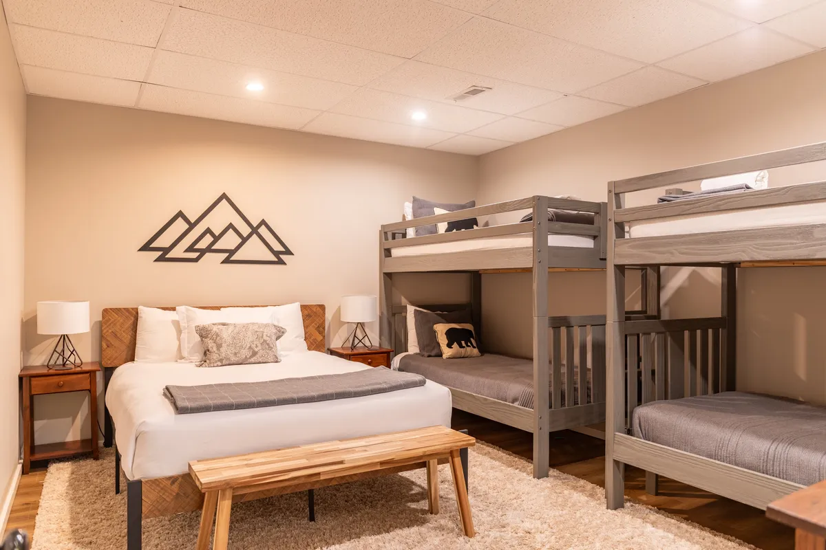 A spacious bedroom featuring a queen bed and two sets of twin bunk beds, accented by mountain-themed wall art and cozy nightstands. The wooden bench and soft lighting create a welcoming, family-friendly atmosphere ideal for kids or groups.