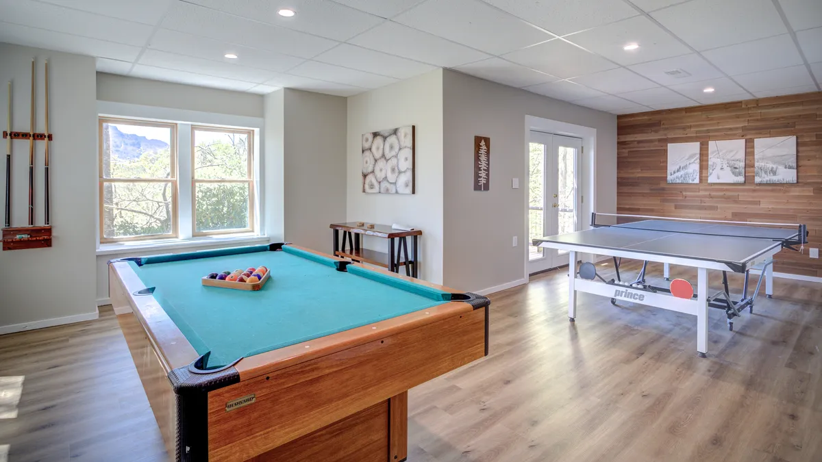 A spacious recreation room featuring both a pool table and ping pong table, with large windows letting in natural light and a wooden accent wall adding warmth to the space. The setup clearly invites friendly competition and group activities, echoing the room’s playful, relaxed atmosphere.